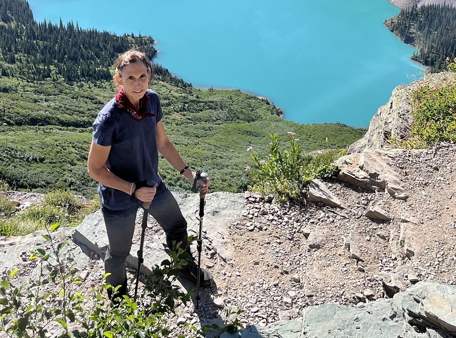 Woman hiker with pole standing above blue lake in mountains.