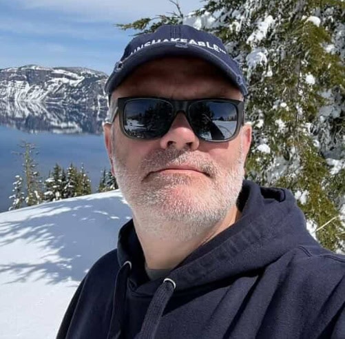 A man with sunglasses and a cap in front of a mounatin scene.