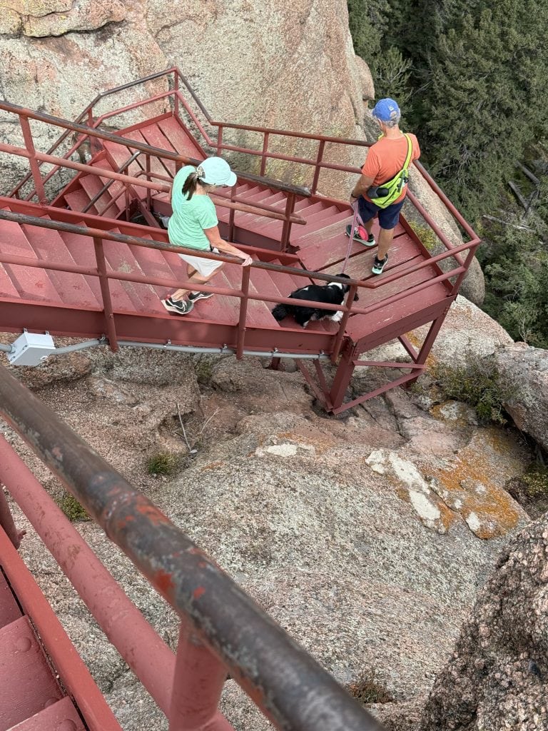 Two hikers and a dog climb down a red metal staircase.